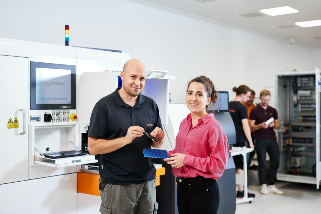 Ein Ingenieur und eine Studentin stecken lachend die Köpfe über einer Solarzelle zusammen. Diese soll mit einer Lasermaschine getrennt werden.